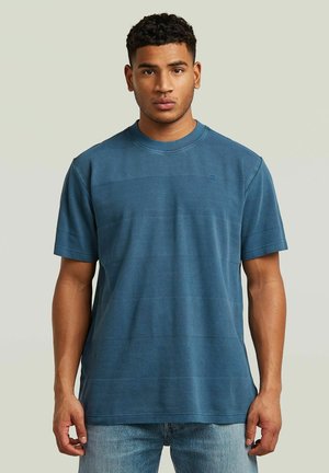 Man met kort krullend haar die een effen blauw shirt met korte mouwen en lichtblauwe spijkerbroek draagt, staat tegen een lichte achtergrond.