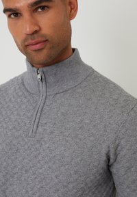 Světle šedý pletený svetr s vysokým límcem, polovičním zipem a texturovaným geometrickým vzorem na látce.