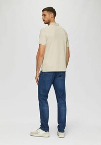 Beige Polohemd mit kurzen Ärmeln und Schlitzen hinten, kombiniert mit dunkelblauen Jeans. Das Model trägt hellfarbene Sneakers und steht mit dem Rücken zur Kamera.