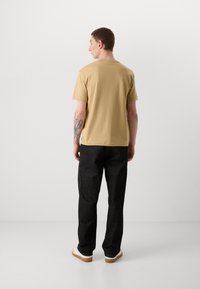 Tan bomull t-shirt med rund halsringning, korta ärmar, i kombination med svarta texturerade byxor och vita sneakers, sedd bakifrån.