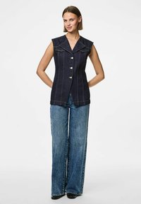 Jeansväst i mörkblått med krage, utan ärmar, frontknäppning och synliga sömdetaljer, kombinerad med vida jeans.