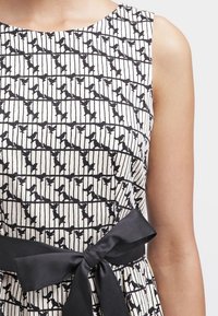 Robe sans manches noir et blanc avec un motif d'oiseaux et de rayures, ornée d'un nœud en satin noir à la taille et d'une texture lisse.