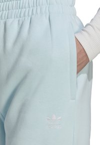 Osoba ubrana w jasnoniebieskie spodnie dresowe Adidas z elastycznym pasem, ręka w bocznej kieszeni, widoczne białe logo Adidas.