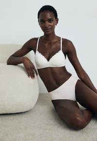 Hvit blonde-bh med bølgende kanter og tynne stropper, parret med en lyserosa sømløs high-waist brief. Myke teksturer og nøytrale farger.