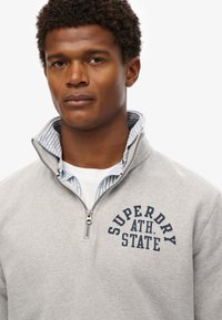 Sudadera gris con cremallera de cuarto de longitud y un logo en azul "SUPERDRY ATH. STATE". Cuello interior a rayas azul y blanco. Tejido de mezcla de algodón.