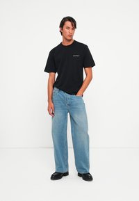 Mann som står med hånden i lommen iført en svart Belstaff t-skjorte, løse lys blå jeans og svarte sko mot en hvit bakgrunn.