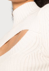 Cremfarbener gerippter Stricktop mit hohem Kragen und Ausschnittdetails an den Schultern. Weiche Textur mit vertikalen Linien und figurbetontem Design.