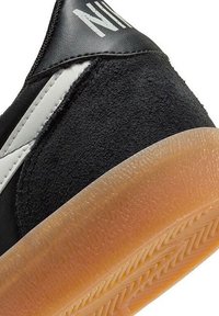 Zapatilla negra con parte superior texturizada, materiales de ante y cuero liso, con acentos blancos, suela de goma y logotipo distintivo en el talón.