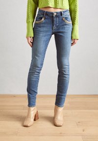 Jeans ajustados azul claro con bolsillos de costura en contraste, combinados con un suéter corto verde brillante y botines de textura beige hasta el tobillo.