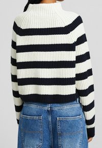 Pletený svetr s námořnickými a krémovými vodorovnými pruhy, s vysokým límcem a žebrovanou strukturou. V kombinaci s džínami vysokého pasu.