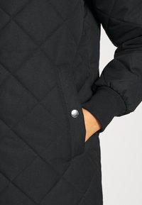 Manteau noir matelassé avec une poche à boutons-pression, des poignets élastiques et une texture de tissu lisse. Des coutures détaillées forment des motifs en losanges.