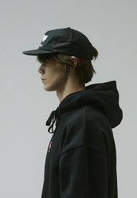 Black baseball cap with mesh panels, featuring a front logo. Worn with a black hooded sweatshirt, displaying a red accent near the neck.