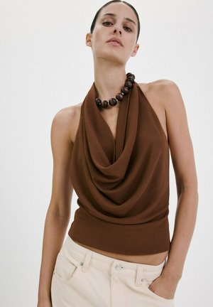 Femme portant un haut marron drapé sans manches à col halter, un pantalon crème et un collier épais de perles en bois foncé, posant avec une main dans la poche.
