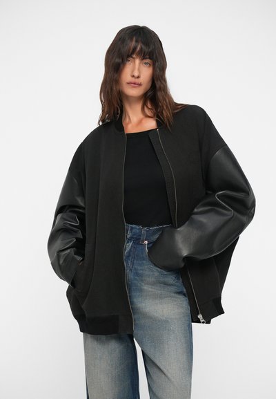 Black bomber jacket with a zip front, featuring oversized fabric body and shiny leather sleeves, paired with faded blue jeans.