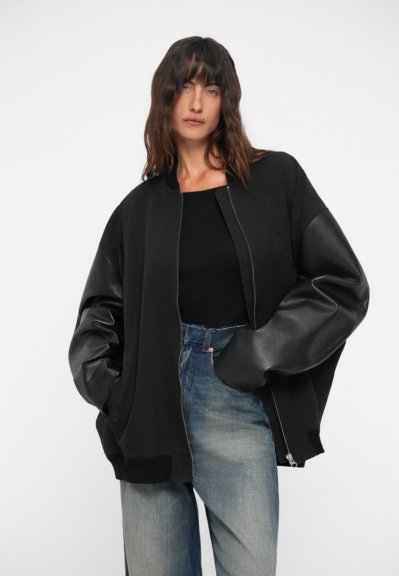 Veste bombardier noire avec fermeture éclair à l'avant, présentant un corps en tissu oversize et des manches en cuir brillant, associée à un jean bleu délavé.