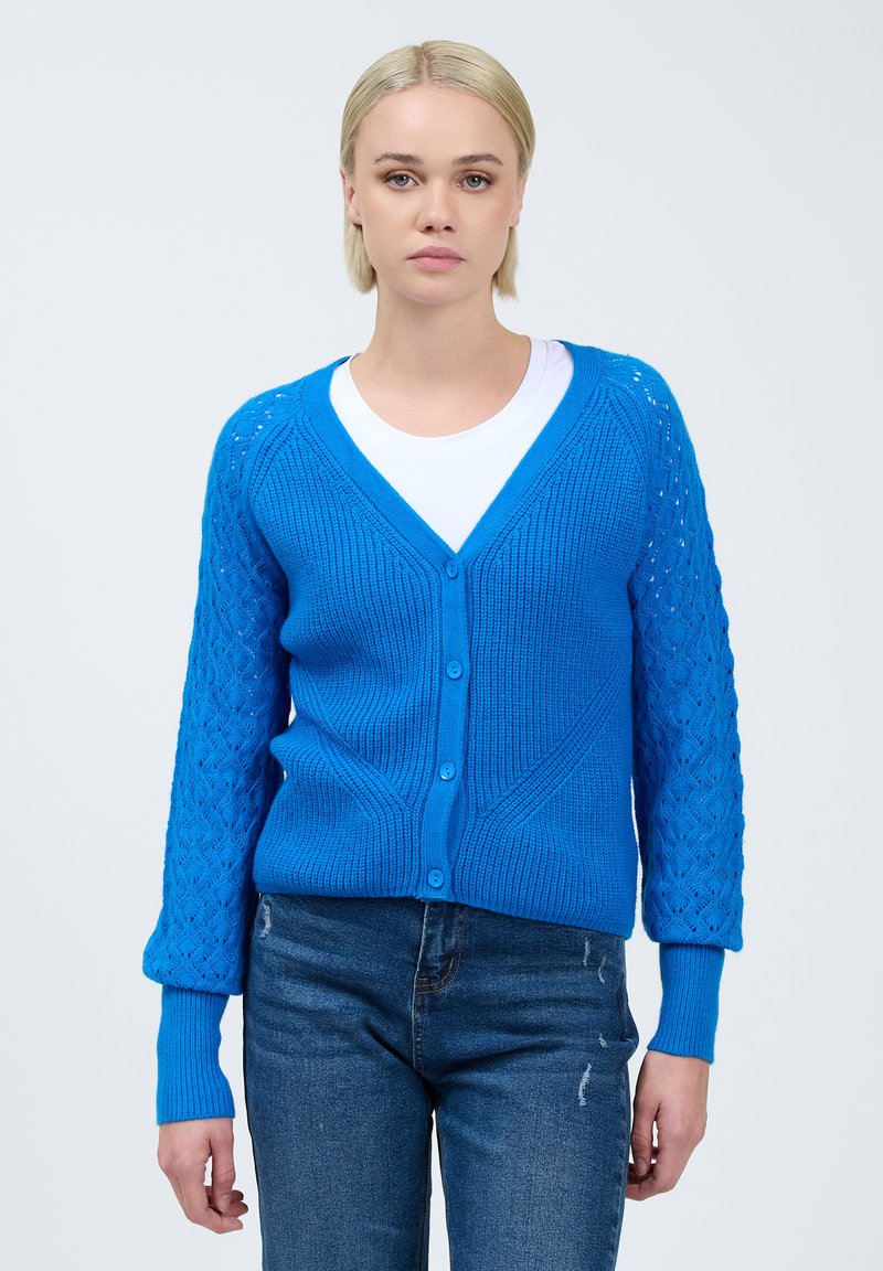 Blau gestrickte Strickjacke mit V-Ausschnitt und Knopfverschluss. Hat strukturierte Ärmel mit einem dekorativen Muster. Kombiniert mit einem weißen Hemd und Jeans.