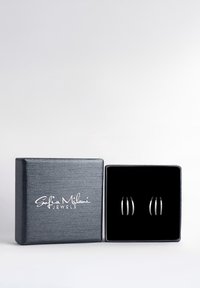 Silver ringörhängen uppvisas i en svart sammetsfodrad låda. Lådan har en texturerad grå utsida och vit logotyptext.