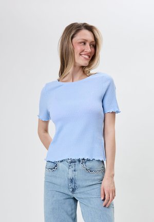 Kvinde iført en lyseblå ribbet kortærmet top og lyseblå jeans med bølget hvid blonderkant på lommerne, smiler og kigger mod højre.