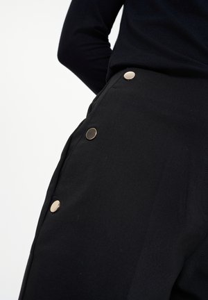 Čierne nohavice s hladkou textúrou, s kovovými gombíkmi v zlatom tóne pozdĺž strany. Zakázkový dizajn s čistou, modernou siluetou.