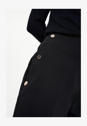 Čierne nohavice s hladkou textúrou, s kovovými gombíkmi v zlatom tóne pozdĺž strany. Zakázkový dizajn s čistou, modernou siluetou.