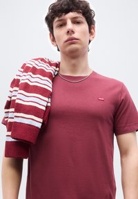 Νεαρός άνδρας που φοράει μαύρη κόκκινη μπλούζα Levi's και ασημένιο αλυσίδα, κρατώντας ένα ριγέ πλεκτό πουλόβερ σε κόκκινο, λευκό και μπλε χρώμα στον ώμο του.