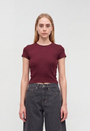 Jeune femme aux cheveux blonds raides portant un haut bordeaux ajusté à manches courtes et un jean large gris foncé, debout devant un fond uni.