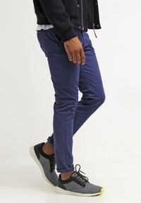Navy blue slim-fit trousers made of stretch material, featuring side pockets and ankle cuffs, paired with black and gray athletic shoes.