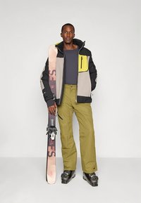 Smučarska jakna s črnimi, sivimi in rumenimi paneli, združena z olivno zelenimi hlačami. Model drži smuči, kar prikazuje zimsko športno opremo.