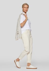 Weißes Poloshirt, beige Hose und weiße Sneaker mit hellbraunen Sohlen. Grauer strukturierter Pullover lässig über die Schulter gelegt. Minimalistisches Design.