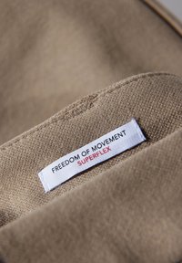 Brązowa tkanina o teksturowanej powierzchni. Biała metka z napisem "FREEDOM OF MOVEMENT" i "SUPERFLEX" w czerwonych i czarnych literach.