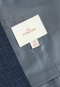 Étiquette de vêtement sur une doublure grise indiquant « NEXT SIGNATURE » avec la taille « 40R » et « Made in Vietnam » sur un fond en tissu bleu à motifs.