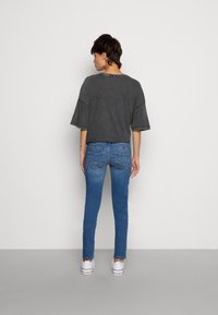 Camisa cinza oversized com mangas curtas, combinada com jeans skinny azuis. Ténis brancos são visíveis. O fundo é branco liso.