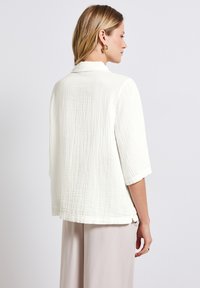 Vit, texturerad blus med en lös passform, trekvartsärmar och subtila fålldetaljer, matchad med ljusa byxor.