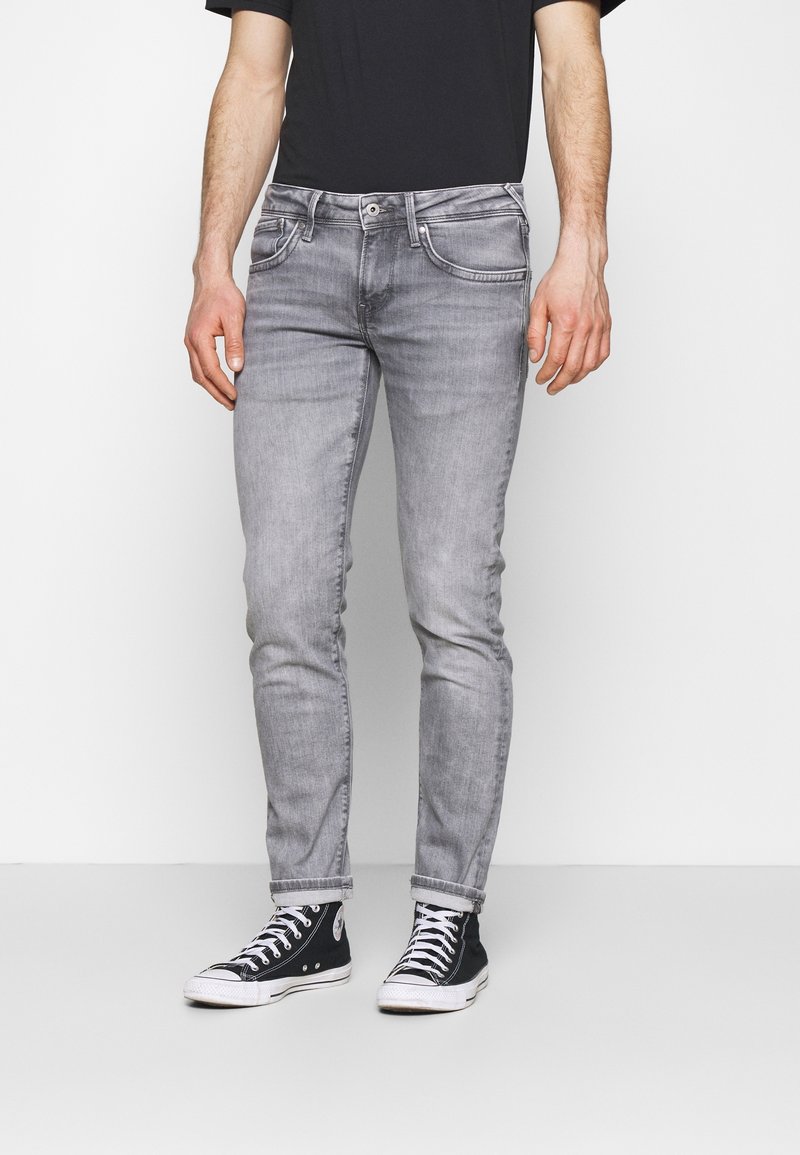 Jean slim gris à texture lisse, avec cinq poches et des détails de coutures discrètes, associé à des baskets montantes noires.