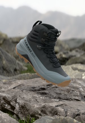 Chaussure de randonnée Mammut noire et grise avec semelle beige en l'air au-dessus d'un terrain rocheux, arrière-plan montagne flou.