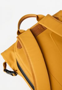 Zaino in tessuto giallo con dettagli in pelle marrone, dotato di spallacci imbottiti, maniglia superiore e tasca laterale con zip per ulteriore spazio di archiviazione.