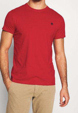 Rotes Baumwoll-T-Shirt mit Rundhalsausschnitt und kurzen Ärmeln, mit einem kleinen schwarzen Logo auf der Brust. Dazu khakifarbene Hosen kombiniert.