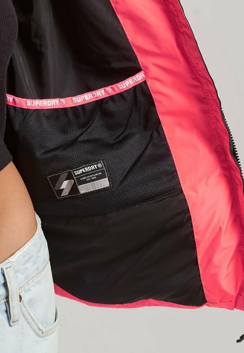 Giacca rossa aperta che mostra la fodera in rete nera con etichetta del logo Superdry e nastro marchiato all'interno, indossata con jeans in denim azzurri.