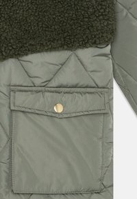 Olivgrön vadderad jacka med en fluffig mörkgrön krage och en ficka med lock som har en snäpplås i guld. Strukturerad tyg och geometrisk sömnad.