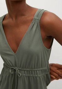 Zielona, bezrękawnikowa sukienka z głębokim dekoltem w serek i zebranymi ramiączkami. Posiada dopasowaną talię z regulowanym sznurkiem i miękką, teksturowaną tkaninę.