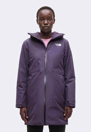 Kobieta w fioletowej bluzie North Face na zamek błyskawiczny z kapturem, stojąca przodem do jasnego, jednolitego tła.