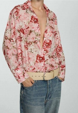 Persoon draagt een losse roze blouse met bloemenpatroon en een diepe halslijn, ingestopt in een spijkerbroek met een brede beige riem, handen in de zakken.