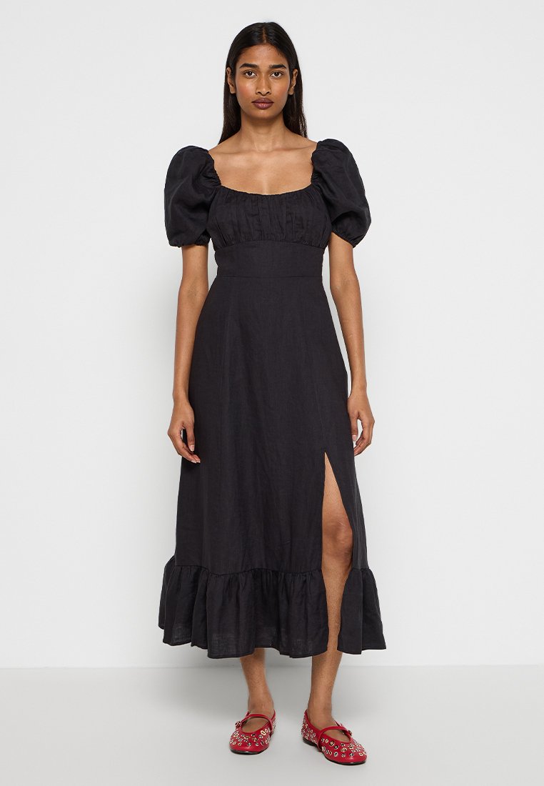 Robe midi en lin noire avec des manches bouffantes, décolleté carré, une fente sur le côté et un ourlet à volants. Associée à des chaussures plates rouges.