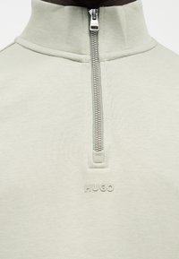 Ανοιχτό γκρι φούτερ με τετραγωνικό φερμουάρ, γιακά που στέκεται και ανάγλυφο λογότυπο "HUGO" στο κέντρο κάτω από το φερμουάρ.