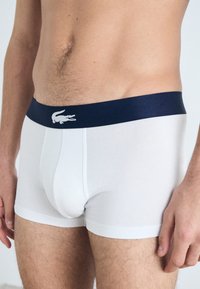 White cotton boxer briefs with a navy elastic waistband, featuring a small white logo. Soft texture, fitted design, and mid-thigh length.