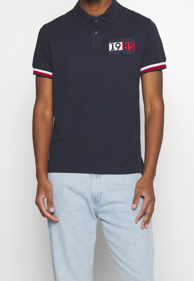 Marineblauw poloshirt met een drieling knoopsluiting, rode en witte gestreepte accenten op de mouwen, en een grafische print met "1985" en "New York."