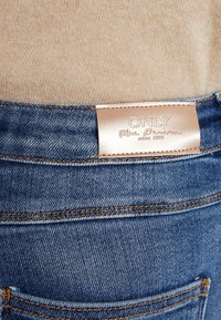 Blå denimjeans med en läderlapp på midjan, med sydda fickor och kontrasterande sömmar i bärnstensfärger.