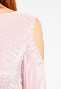 Frau mit roten Haaren, die ein hellrosa Oberteil aus geripptem Stoff mit schulterfreien Ausschnitten vor einem weißen Hintergrund trägt.