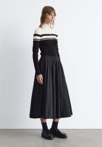 Schwarzer Rock mit einem fließenden Design, kombiniert mit einem enganliegenden schwarz-weiß gestreiften Ripp-Pullover. Das Modell trägt schwarze Schuhe und Socken.