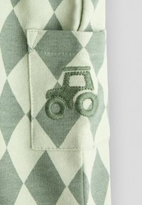 Zaļas un baltas dimanta raksta audums ar mazu kabatiņu, kurā ir izšūts zaļš traktora dizains.